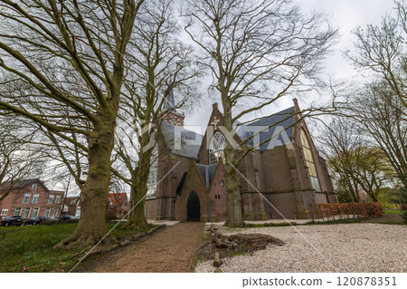 A historic brick church surrounded by tall, bare trees with visible roots. The gothic architecture is highlighted against an overcast sky, creating a serene atmosphere. Church near Amsterdam. 120878351