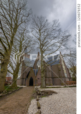 A historic brick church surrounded by tall, bare trees with visible roots. The gothic architecture is highlighted against an overcast sky, creating a serene atmosphere. Church near Amsterdam. 120878352