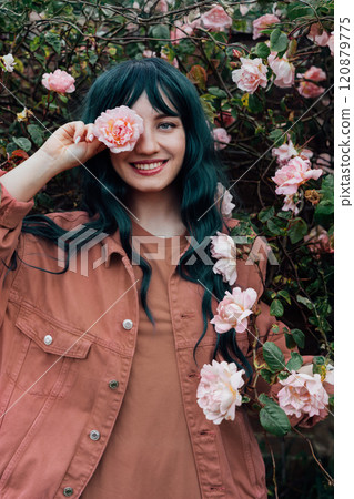 Young hipster woman with color hair in mocha color dress holding flower on blooming roses bush background. Urban street fashion. Fun, positive mood. Trendy fashion outfit. Color of the 2025 year 120879775