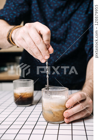 Close-up of hands stirring creamy iced coffee in ribbed glass. 2025 color Mocha Mousse Close-up of hands stirring creamy iced coffee in ribbed glass. 2025 color Mocha Mousse 120879955