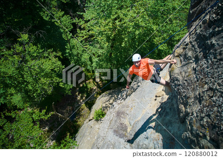 Aerial view of male rock climber ascending rugged limestone cliff with harness and rope for safety. Sportsman climbing on vertical large boulder at Dobvush Rocks in Carpathian mountains, Ukraine. Aerial view of male rock climber ascending rugged limestone cliff with harness and rope for safety. Sportsman climbing on vertical large boulder at Dobvush Rocks in Carpathian mountains, Ukraine. 120880012