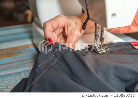 Hands of a eldlerly seamstress sewing a pair of pants on a sewing machine. 120880738
