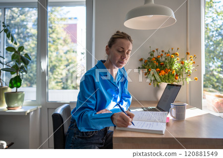 Focused woman comfortable works at home by writing notes in day planner sits at table with laptop. Focused woman comfortable works at home by writing notes in day planner sits at table with laptop. 120881549