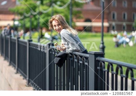 Portrait joyful woman walking in city park with ice cream standing near water fence looks to camera Portrait joyful woman walking in city park with ice cream standing near water fence looks to camera 120881567