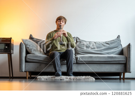 Clarinet player. Man clarinetist playing flute woodwind music instrument sitting on couch at home 120881625
