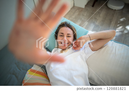 Sleepy middle-aged woman lies on bed smiling and stretching raised one hand close up to camera. 120881821