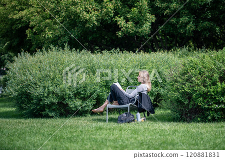 Serene woman deeply engaged in book, sprawled on chair in park. Study and digital detox, city break 120881831