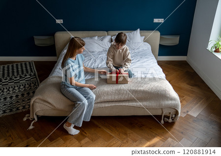 Mother sitting on bed with little boy child opening wrapped gift box 120881914