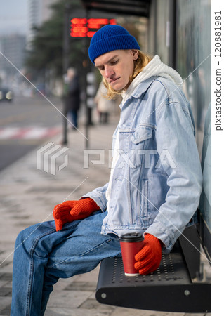 Young man sits on bench at bus stop. Guy with coffee cup calmly waiting for public transport. 120881981