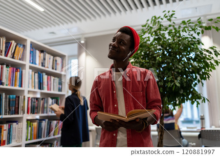 Joyful black student guy stands in university library with book thinks about good job after diploma. Joyful black student guy stands in university library with book thinks about good job after diploma. 120881997