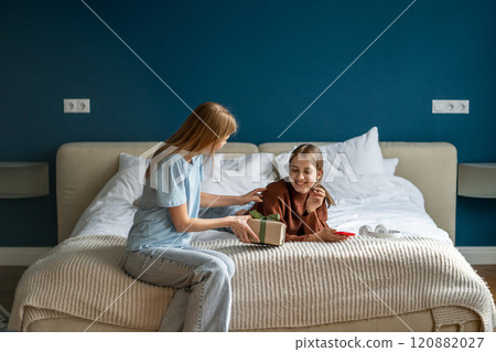 Happy surprised adolescent teen girl receiving birthday present from mother while resting on bed Happy surprised adolescent teen girl receiving birthday present from mother while resting on bed 120882027