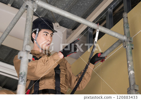 A person checking the safety of scaffolding. A person wearing work clothes and a safety harness says, "All right!". An image of a construction worker or blue collar worker. Please be safe. 120883433