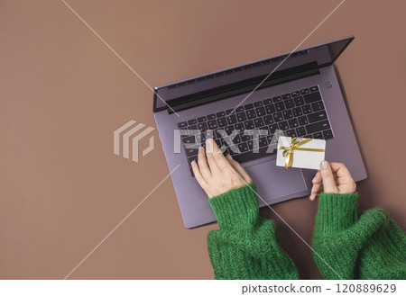 Christmas shopping concept. Flat lie. Women's hands in a green sweater on laptop keyboard with Gift card on a brown mocha background 120889629