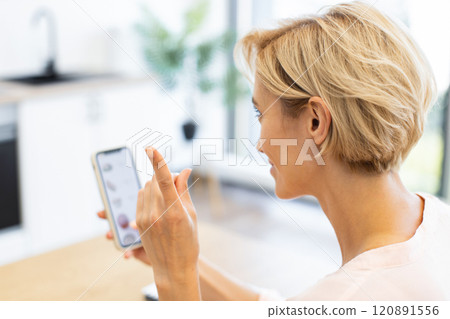 Caucasian woman using smartphone to order food online in bright, modern kitchen. Focused on screen, she appears engaged, utilizing a delivery app. Caucasian woman using smartphone to order food online in bright, modern kitchen. Focused on screen, she appears engaged, utilizing a delivery app. 120891556