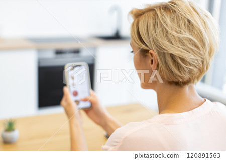 Caucasian woman using smartphone to order food online in modern kitchen setting. Blonde woman with short hair browsing delivery website. Image reflects convenience, technology, and digital lifestyle. Caucasian woman using smartphone to order food online in modern kitchen setting. Blonde woman with short hair browsing delivery website. Image reflects convenience, technology, and digital lifestyle. 120891563
