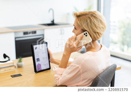 Caucasian woman using smartphone while browsing clothing website on tablet. Sitting at desk in modern kitchen, ordering clothes online with delivery service. 120891570