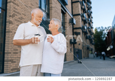 Loving retired couple in casual clothes enjoying coffee standing on city street, smiling and talking, savoring togetherness in urban landscape on sunny day, on blurred background of building exterior. 120892287