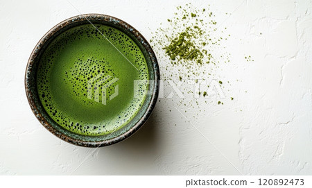 A close-up view of a rustic bowl filled with vibrant green matcha tea, accompanied by a sprinkle of matcha powder on a light textured surface, ideal for healthy beverage promotion. 120892473