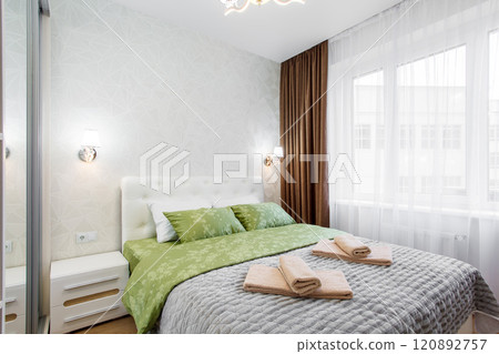 A well-appointed small modern bedroom with a cozy design, featuring a queen-sized bed, bedside tables. A large window with curtains allowing soft daylight. 120892757