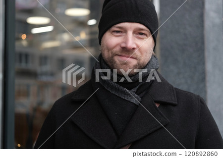 Handsome man wearing black coat and brown leather bag walking down on the city with mobile phone in hand. Stylish bearded middle-aged male using smartphone at urban street. Businessman going on job. 120892860