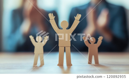 Wooden figures representing success and achievement, with individuals celebrating in the background, symbolizing teamwork and support in a corporate environment. 120892935
