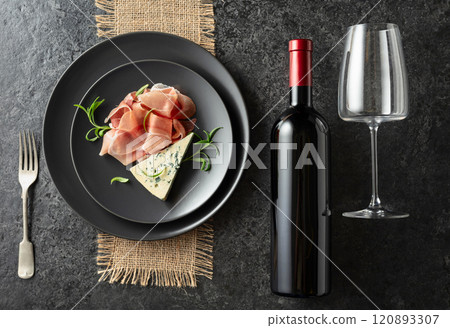 Bottle of red wine with blue cheese, prosciutto, and rosemary on a black table. 120893307