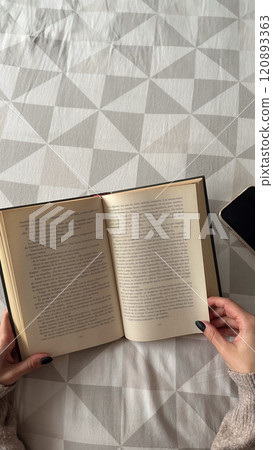 Woman lying on bed, close up shot of female hands holding book and mobile, book day, entertainment 120893363