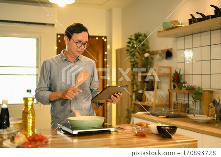 Portrait of senior man following a recipe on digital tablet while cooking in his home kitchen Portrait of senior man following a recipe on digital tablet while cooking in his home kitchen 120893728