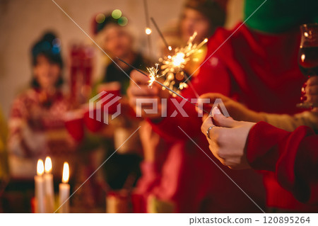 Hands holding sparklers light up festive scene, with blurred friends in colorful winter outfits and red cups celebrating in cozy atmosphere on background. Hands holding sparklers light up festive scene, with blurred friends in colorful winter outfits and red cups celebrating in cozy atmosphere on background. 120895264