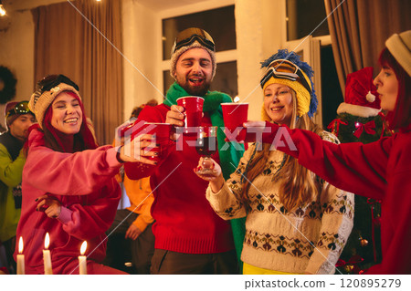Lively group of friends dressed in winter sweaters and ski hats enjoys drinks at festive wooden table, surrounded by holiday-themed decorations. Lively group of friends dressed in winter sweaters and ski hats enjoys drinks at festive wooden table, surrounded by holiday-themed decorations. 120895279