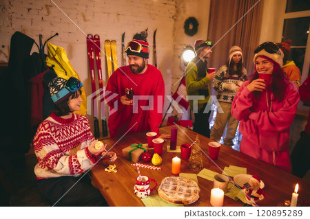 Festive spirit fills room as friends in ski outfits share drinks and laughter around holiday-themed table with candles, gifts, and seasonal treats. 120895289