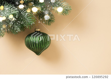Christmas green bauble on fir snowy branch and lights garland on beige background. Xmas greeting card. New Year banner. Copy space. View from above. Christmas green bauble on fir snowy branch and lights garland on beige background. Xmas greeting card. New Year banner. Copy space. View from above. 120895292