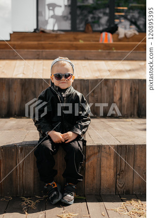 Fashion kid summer in the city stands wood wall. Trendy boy in sunglasses standing on the street at sunset. Fashion kid summer in the city stands wood wall. Trendy boy in sunglasses standing on the street at sunset. 120895395