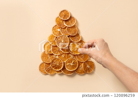 Woman make Eco-friendly creative Christmas tree made of natural dried orange slices on beige background. Food vegan. Happy New Year. View from above. Xmas greeting card. Woman make Eco-friendly creative Christmas tree made of natural dried orange slices on beige background. Food vegan. Happy New Year. View from above. Xmas greeting card. 120896259