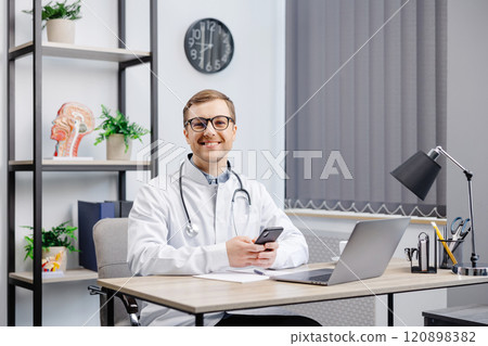 Male doctor medical worker in modern clinic wearing eyeglasses and white coat uniform using cell mobile smartphone apps, sitting at laptop computer. Medicine technologies health care concept. 120898382
