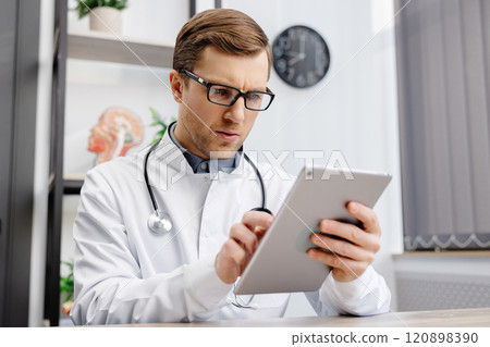 Male Caucasian doctor in white medical uniform filling patient medical history or anamnesis on tablet in office. Doctor consulting client online on tablet in modern clinic, healthcare concept Male Caucasian doctor in white medical uniform filling patient medical history or anamnesis on tablet in office. Doctor consulting client online on tablet in modern clinic, healthcare concept 120898390