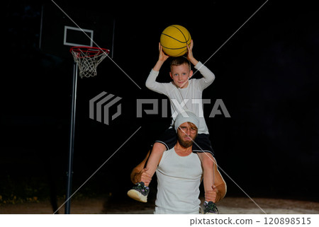 A father and son share a fun moment playing basketball outdoors at night with a hoop in the background 120898515