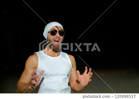 A man wearing a tank top, sunglasses, and a beanie poses confidently at night against a dark background A man wearing a tank top, sunglasses, and a beanie poses confidently at night against a dark background 120898527