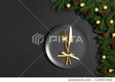 Christmas festive table setting on black background. Black plate, gold cutlery and fir branches with decorations for New Year's Eve dinner. Copy space. Top view, flat lay. 120903468