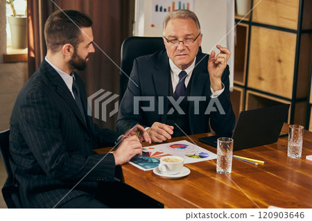 Senior businessman explaining data points to colleague, with charts and laptop on the table during team meeting. Personal professional consultation. Senior businessman explaining data points to colleague, with charts and laptop on the table during team meeting. Personal professional consultation. 120903646