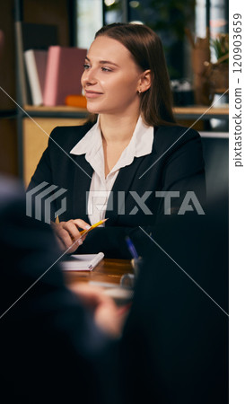 Confident young businesswoman in formal attire attentively listening during professional meeting, holding pen and notebook. 120903659