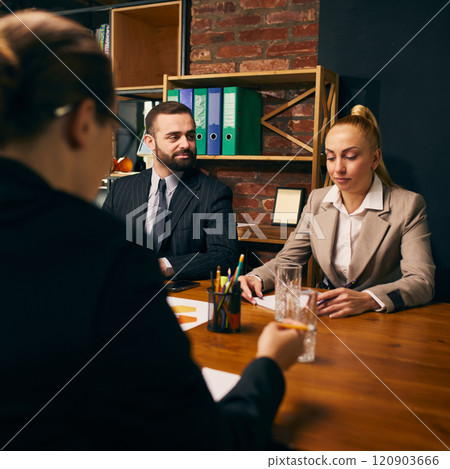 Two professionals in formal attire actively participate in business meeting, with office shelves in background. Professional collaboration Two professionals in formal attire actively participate in business meeting, with office shelves in background. Professional collaboration 120903666