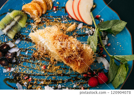 Elegant dessert with kunafa and fresh fruit slices on a blue plate. A creative presentation of Middle Eastern sweets with vibrant garnishes. Elegant dessert with kunafa and fresh fruit slices on a blue plate. A creative presentation of Middle Eastern sweets with vibrant garnishes. 120904078