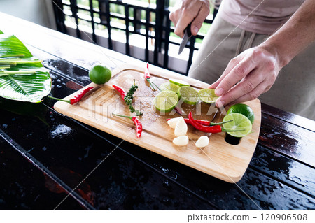 Fresh limes are being sliced alongside vibrant red chilies and garlic cloves on a wooden cutting board. The tropical kitchen has a lively atmosphere with natural light. 120906508