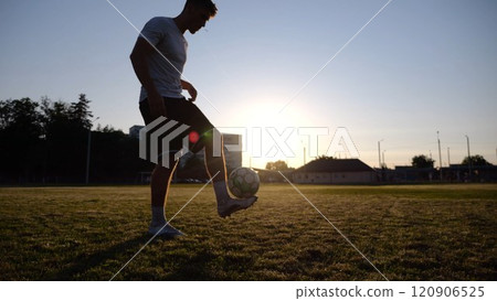 Professional footballer juggling soccer ball on stadium at sunset. Young man kicking ball at green field. Sportsman practicing tricks at meadow with sunlight at background. Freestyle football concept 120906525