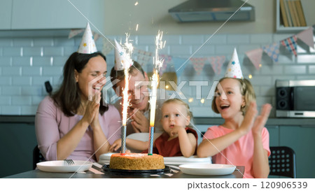 Happy family of four gathers around a baby in a high chair, clapping and cheering as they watch sparkling ice fountains on the cake 120906539