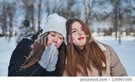Two schoolgirls standing in a snowy park, warming hands during winter snowfall, bundled up in warm clothing. Happy and cheerful, showcasing friendship in the cold 120907309