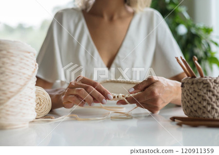 A young girl in a white dress sits at a table and knits a basket from beige yarn. 120911359