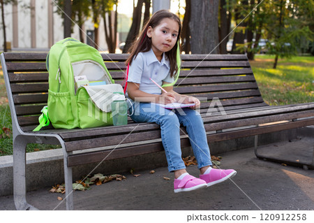 Young girl writing in notebook on park bench with backpack Young girl writing in notebook on park bench with backpack 120912258