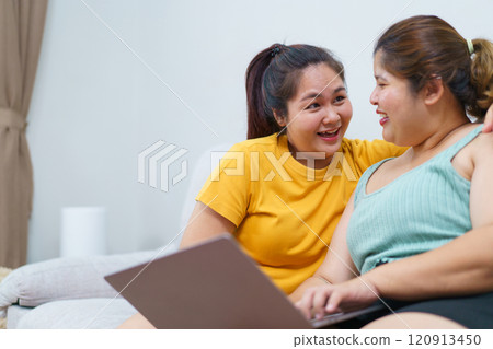 Happy Asian women friend relaxing in living room and drink a cup of tea together. Happy - delighted two Asian oversized women talking and watching a movie on laptop or notebook computer. Happy Asian women friend relaxing in living room and drink a cup of tea together. Happy - delighted two Asian oversized women talking and watching a movie on laptop or notebook computer. 120913450